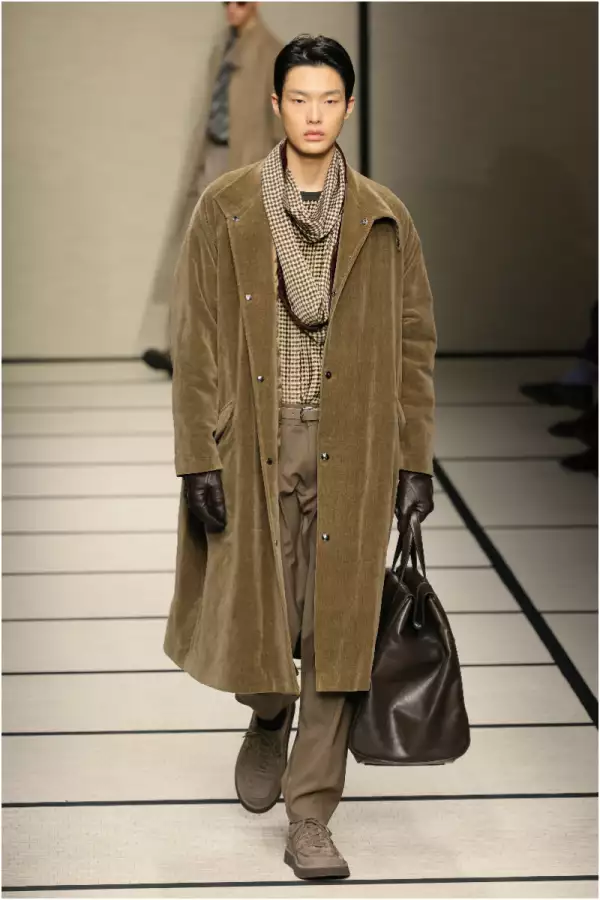 Foto de un modelo de Armani por la pasarela con un abrigo café debajo de las rodillas, una bufanda en tonos tierra y un bolso grande en la mano.
