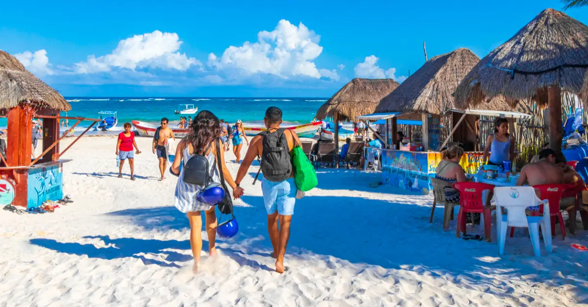 Una pareja camina de mano en una playa con arena blanca y mar y cielo azul. Palapas y sillas donde hay personas sentadas en sillas.jpg