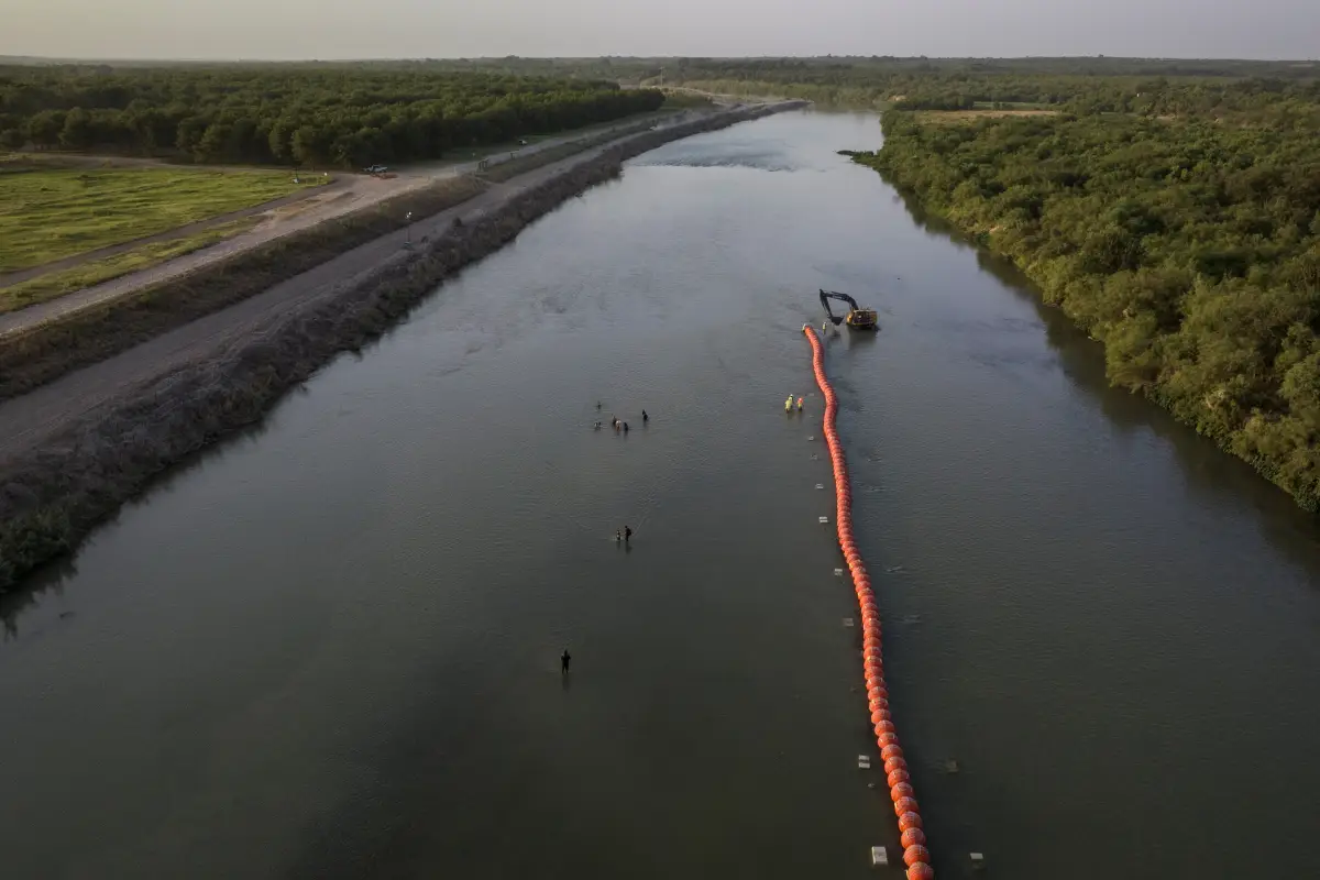 El huerto de nueces de Heavenly Farms se ve a la izquierda mientras las familias migrantes navegan por el río Río Grande pasando por trabajadores y boyas mientras buscan un punto de entrada a los Estados Unidos desde México en Eagle Pass, Texas, EE. UU., el 27 de julio de 2023.
