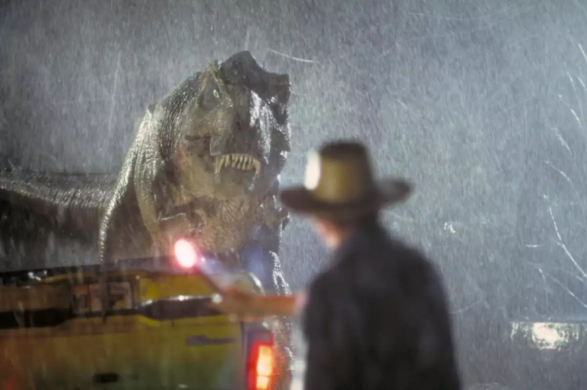Foto de una escena de la película 'Jurassic Park' de 1993 con el tiranosaurio rex a lo lejos con expresión amenazante bajo la lluvia.