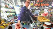 Una mujer de cabello corto y oscuro vestida con una blusa morada detrás de un mostrador cobra refrescos a un cliente.