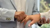 Una mano de hombre sostiene otra y le coloca un anillo en un dedo. Uno viste traje azul y el otro, blanco.jpg