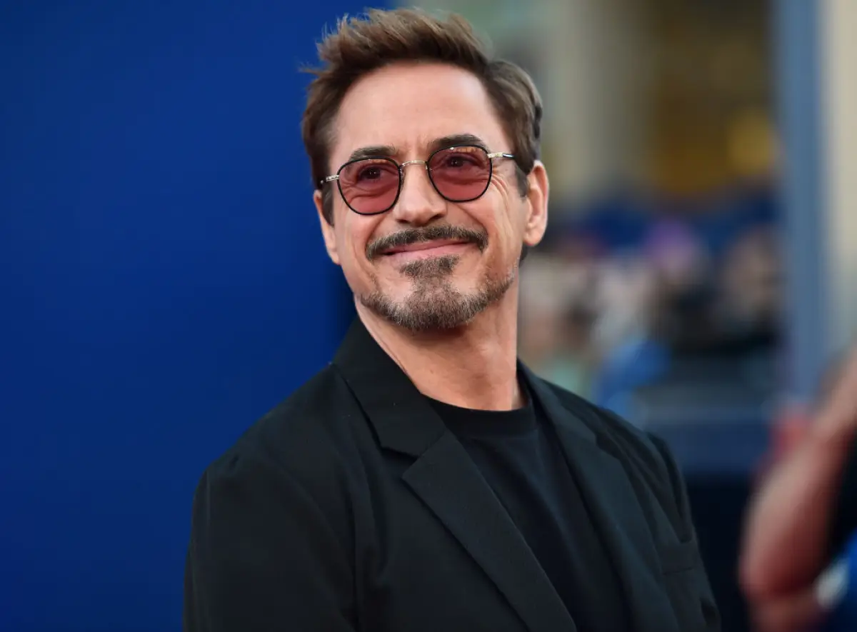 Foto de Robert Downey Jr sonriendo, viste saco y playera negra y lleva lentes oscuros en un tono rojizo.