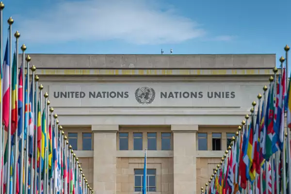 El palacio de las Naciones Unidas en Ginebra, se ve el edificio con el logo de naciones unidas al centro al igual que su bandera azul. A los lados del jardín (parcialmente visible) se encuentran las banderas de todas las Naciones que forman parte.