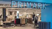 Entrada de una tienda Primark, en la que se muestran aparadores con ropa y zapatos de colores neutros como gris y nude. La fachada es color azul, con el nombre de la cadena en el mismo color en la parte de arriba de la entrada.

