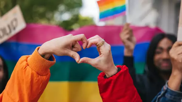 Genderqueer and non-binary friends making a heart shape against a rainbow flag
