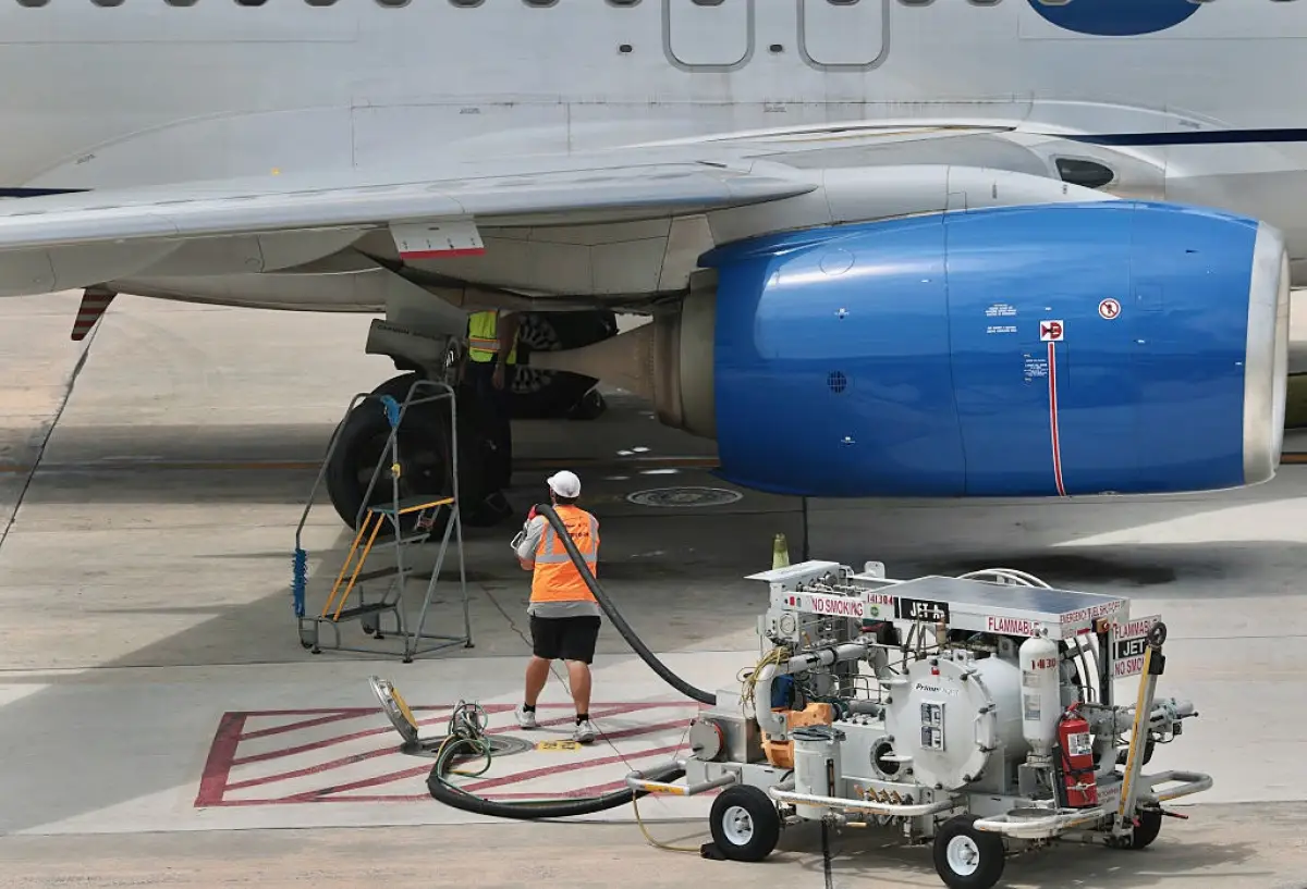 Un avión comercial estacionado en la plataforma de un aeropuerto mientras trabajadores realizan el abastecimiento de turbosina mediante una manguera conectada a un equipo especializado. La operación se lleva a cabo bajo el ala de la aeronave, junto al motor y el tren de aterrizaje, con equipos y señalizaciones de seguridad para prevenir riesgos durante el suministro de combustible.
