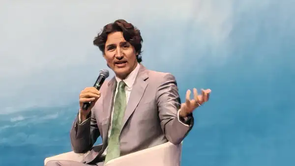 Justin Trudeau, exprimer ministro de Canadá, habla durante un evento sentado en un sillón blanco, con traje gris claro, camisa blanca y corbata verde. Sostiene un micrófono y gesticula con la mano izquierda frente a un fondo azul.