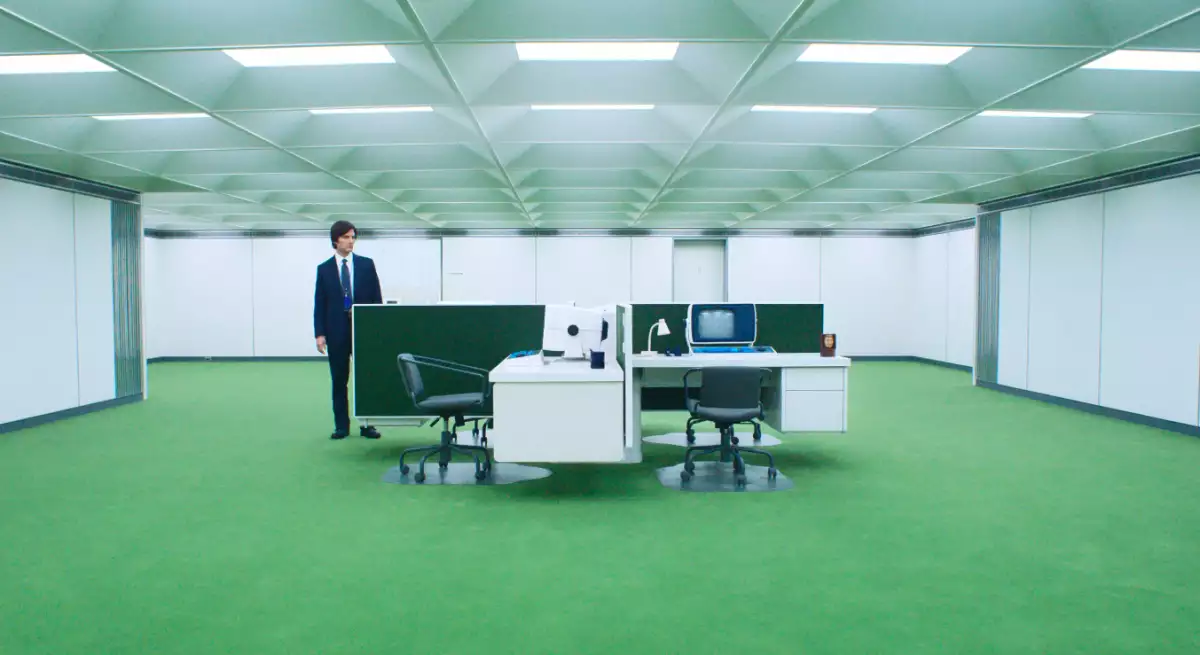 Foto de una escena de la serie Severance donde se ve a Mark (Adam Scott) parado en un enorme cuarto de paredes y luces blancas y en el centro hay escritorios de trabajo.