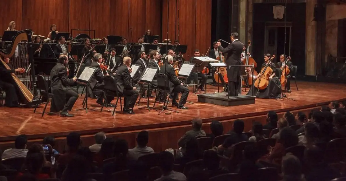Músicos con violines y partituras con un director de orquesta en un teatro de color café frente a personas sentadasjpg
