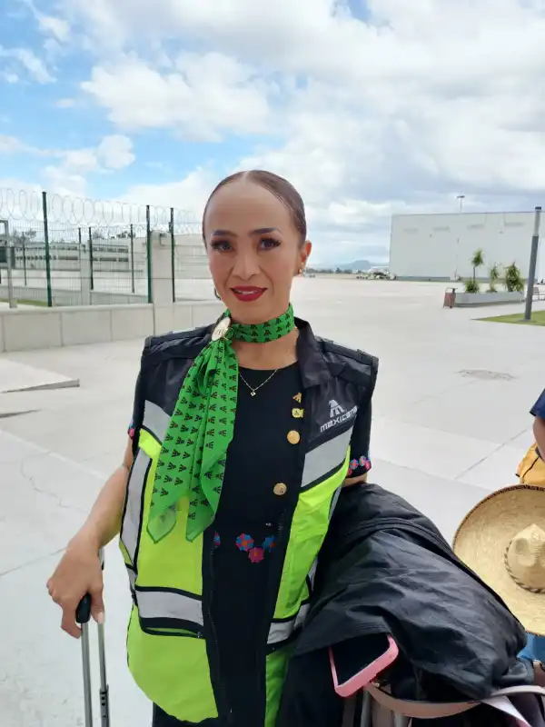Una mujer con falta y chaleco azul, chaleco verde y pelo recogido, agarrando una maleta a cielo abierto.jpeg