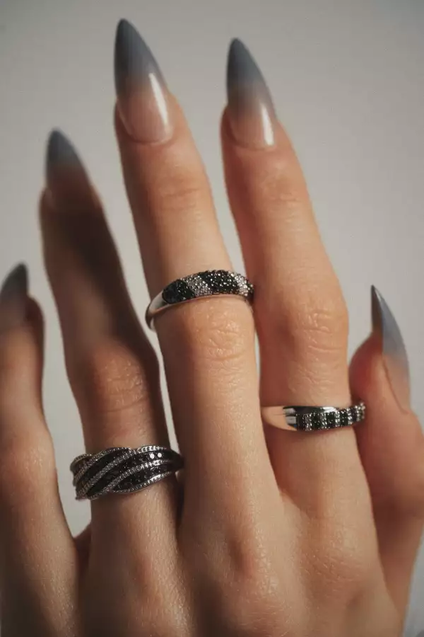 Foto de una mano con uñas postizas y entre los dedos luce tres anillos de Bizzarro de diamante negro y oro blanco