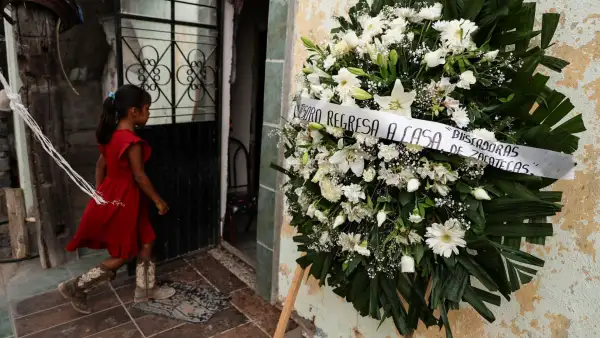 Niña con vestido rojo y botas camina frente a una casa junto a una corona funeraria con un mensaje de familiares de personas desaparecidas.