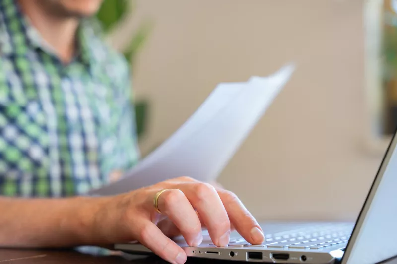 Closeup Of Hand Typing On Laptop Keyboard Holding Paper Documents Remote Work