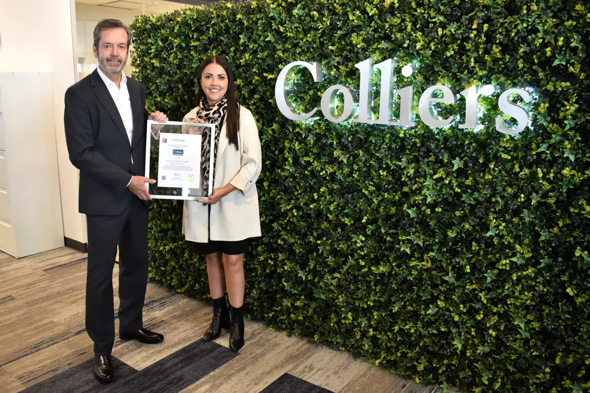Hombre y mujer sostienen un reconocimiento, detrás se observa un muro verde con la leyenda Colliers,