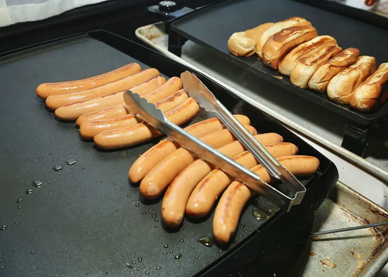 Una plancha industrial muestra varias salchichas dorándose, con pinzas metálicas colocadas sobre ellas. Al fondo, una segunda superficie caliente sostiene varios panes para hot dogs, algunos tostados por los lados. La escena corresponde a la preparación de salchichas para su servicio, con gotas de aceite visibles sobre la plancha.