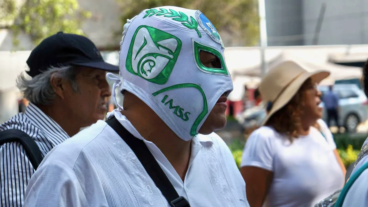 Un hombre con máscara de luchador blanca con verde con letras que dicen IMSS. Un hombre mayor y una mujer con sombrero.jpg