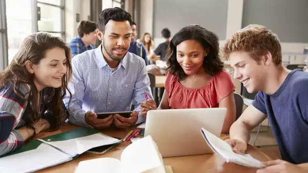 High School Students Using Laptops And Digital Tablets Working With Male Teacher At Desk