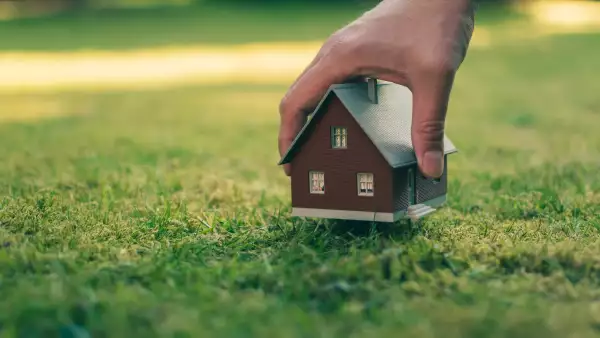 Concept of selling a house. A hand is holding a model house above green meadow.