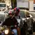 Dos personas viajan en una moto, con otros vehículos de fondo.