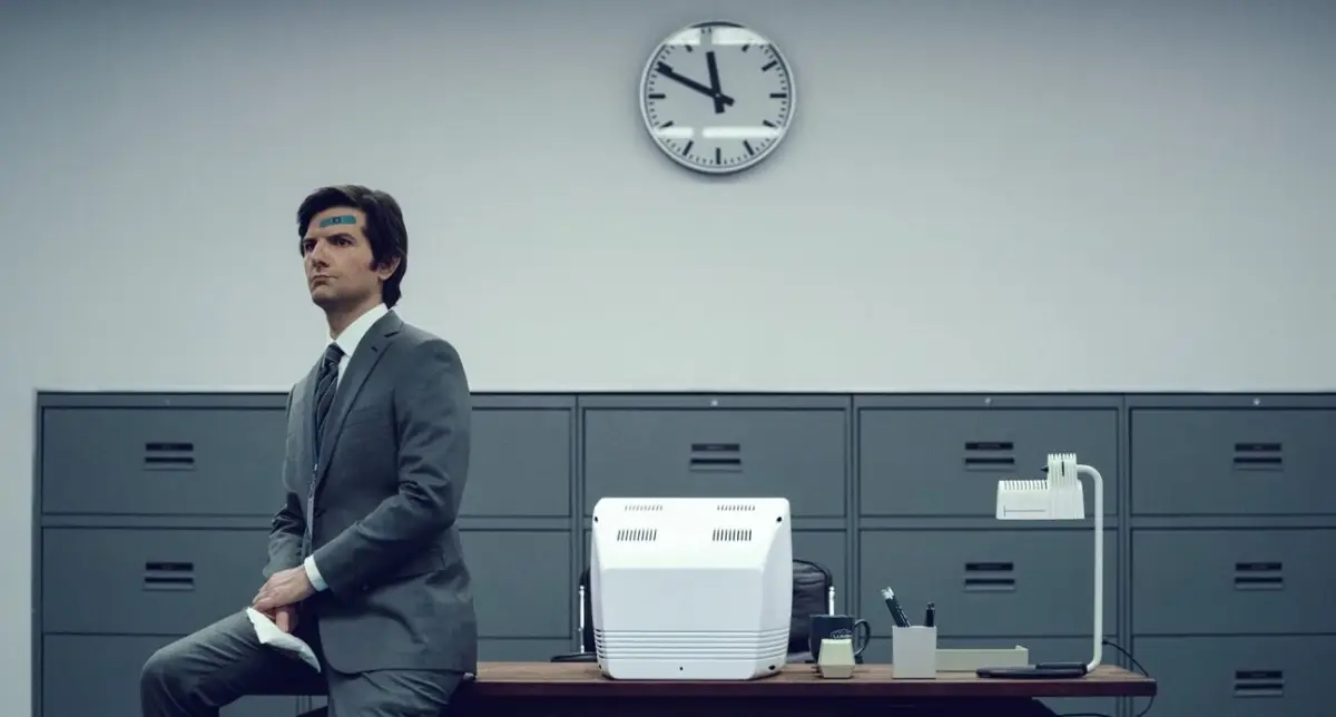 Foto de Adam Scott como Mark, personaje de 'Severance' sentado en la esquina de un escritorio vestido de gris en una oficina llena de archiveros grises.