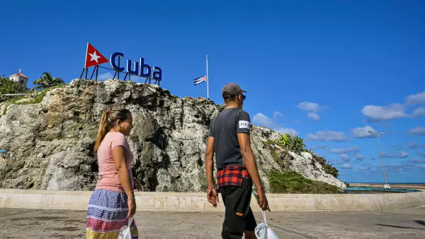 Los cubanos caminan por un cartel de Cuba y una bandera nacional a media asta en La Habana el 5 de enero de 2026. La Habana declaró dos días de luto nacional a partir del 5 de enero después de que un total de 32 cubanos fueran asesinados durante el ataque estadounidense a Caracas que culminó con la captura del presidente venezolano Nicolás Maduro.