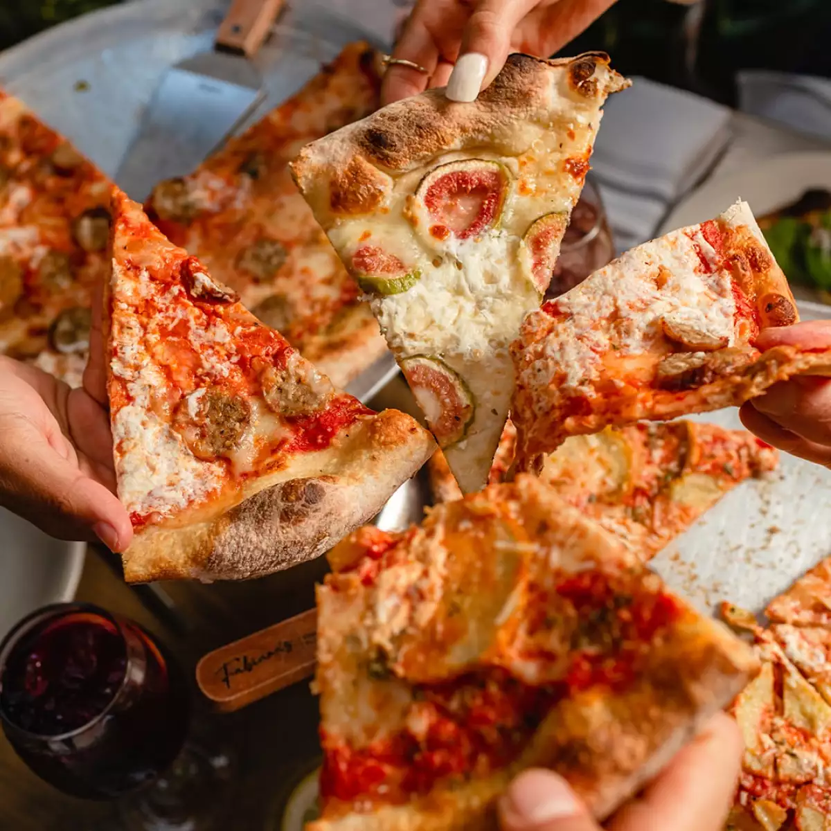 Foto de cuatro manos, cada una con un triángulo de pizza como "brindando" para mostrar las opciones del restaurante