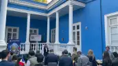 Ventanales y columnas de una casa azul con blanco. Personas sentadas en el exterior frente a una mujer con micrófono.jpg